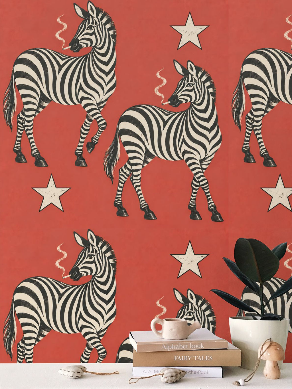 Red wallpaper featuring a repeating pattern of black and white zebra illustrations and cream stars, shown behind a tabletop styled with books, a mug, a plant, and small decorative objects.
